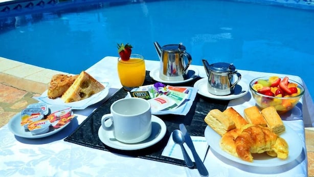 Desayuno junto a una piscina con tostadas, croissants, fruta, una cafetera y un vaso de zumo de naranja.
