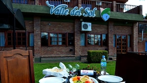 Un montaje para comer al aire libre con una mesa lista para dos personas, una botella de agua y una de vino. Al fondo, se observa un edificio con un letrero que dice "Valle del Sol".