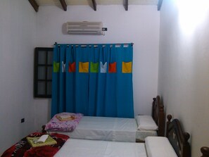 A small, simple room with two beds, a window with a curtain, and an air conditioning unit on the wall.