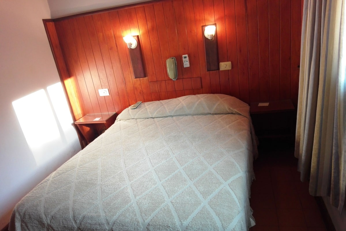 A hotel room with a wooden headboard, two wall-mounted lights, a bedside table, and a bed with a patterned bedspread.