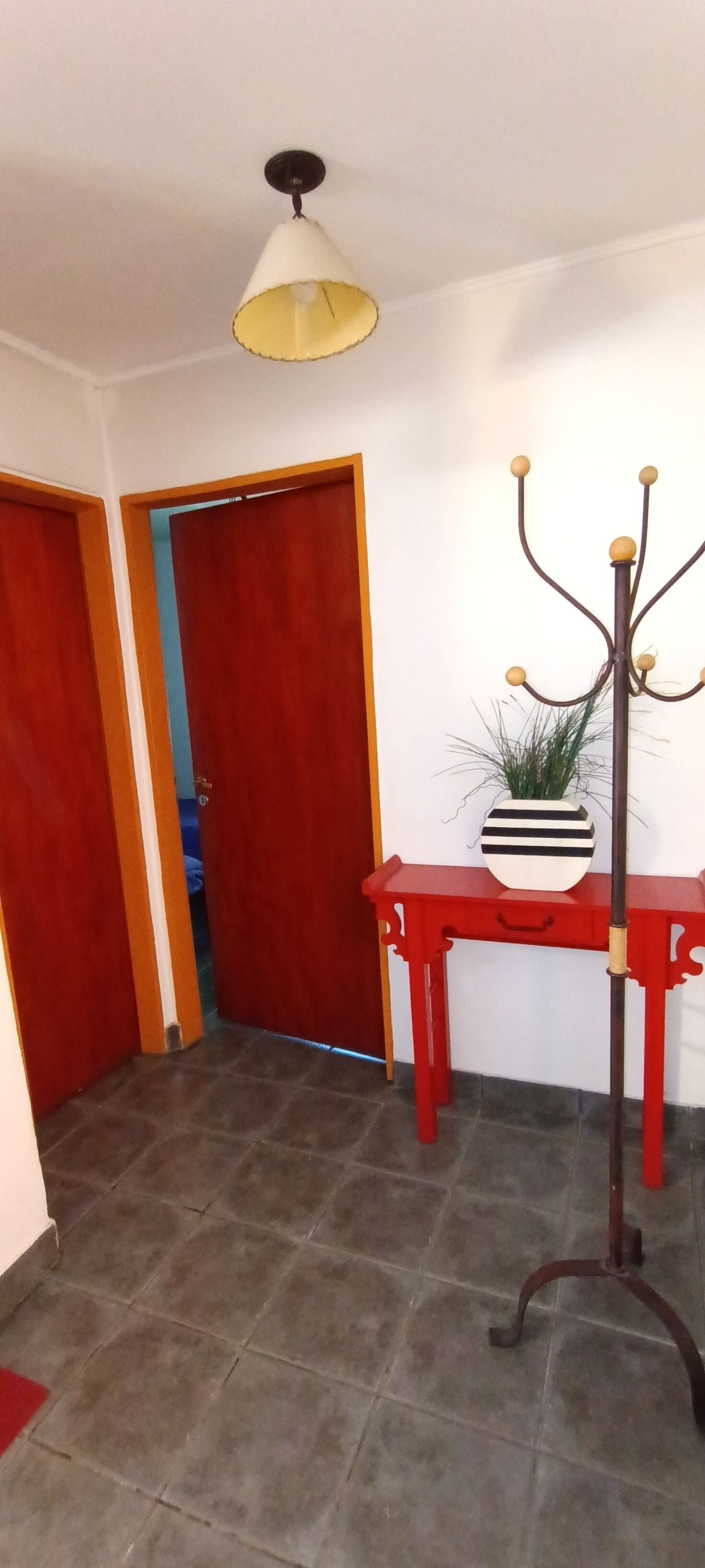 Un couloir avec une porte rouge, une table rouge ornée d’une plante et un luminaire suspendu.