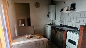 Cocina compacta con estufa, lavamanos y un refrigerador pequeño. Hay una zona de comedor con una silla y una mesa, y un ventanal que proporciona luz natural.