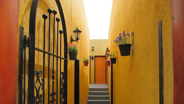 Un estrecho callejón con una puerta de madera al final, flanqueado por paredes amarillas y rejas de metal negro.