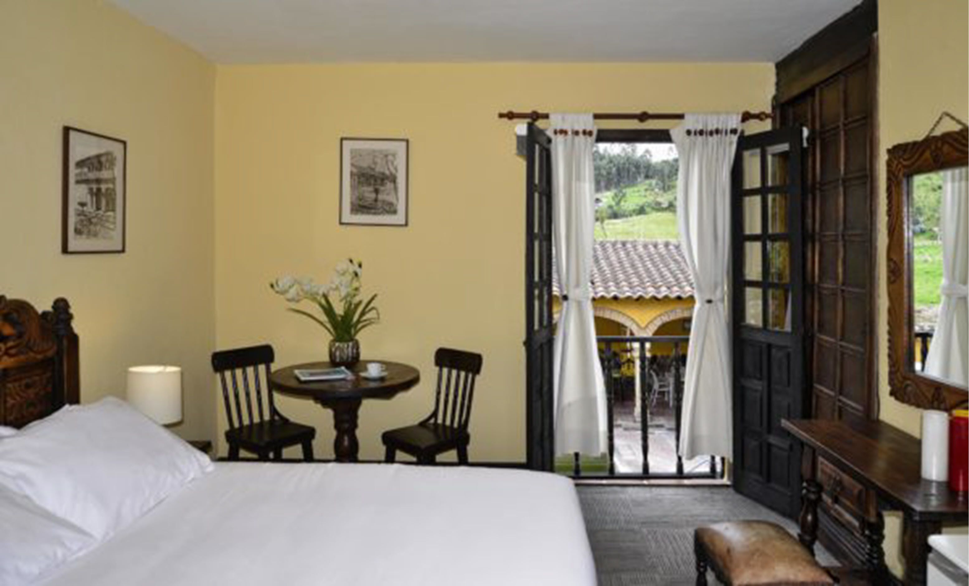 Una habitación de hotel con una cama, una mesa con sillas, un florero y vistas al exterior a través de una puerta abierta.