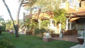 A courtyard with a brick building, palm trees, and a grassy area. - Del Pomar Hotel Boutique (Resistencia)