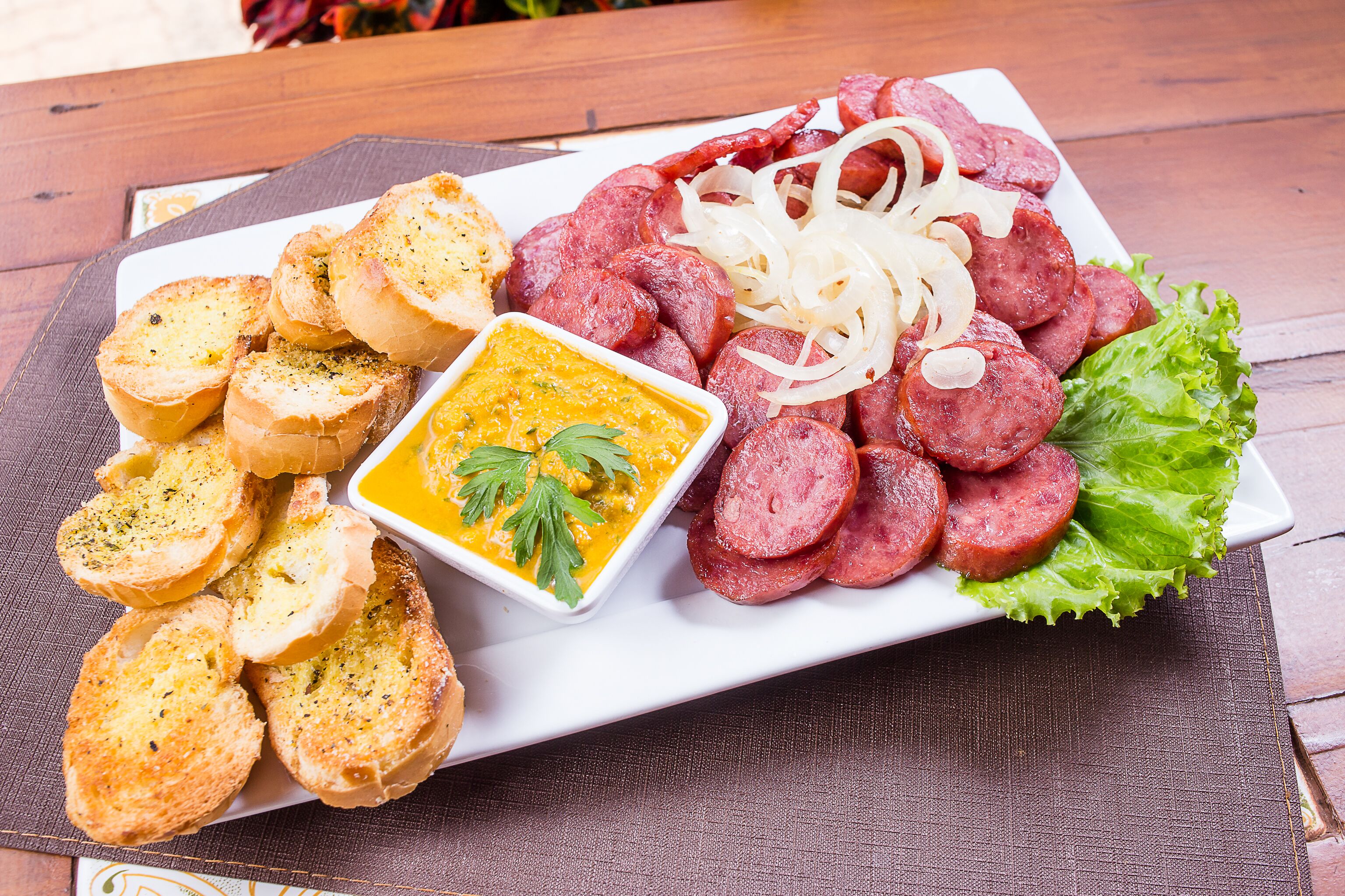Un plat de tall de botifarra, pa torrat i un bol petit de salsa damunt un taulell de fusta.