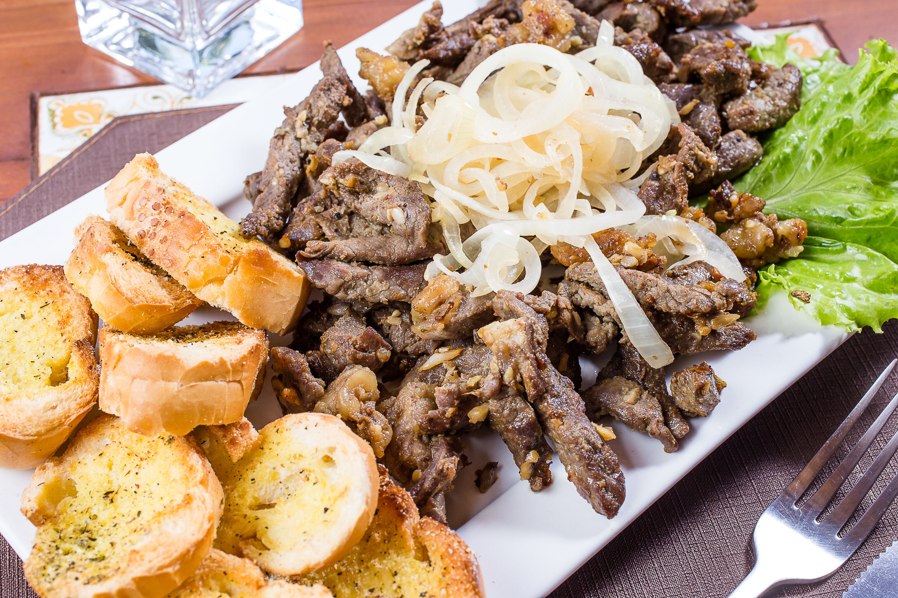 A plate of grilled meat with onions and lettuce, accompanied by toasted bread slices.