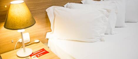 Una cama bien hecha con sábanas blancas, una lámpara de noche, un control remoto y una señal roja sobre la mesita de noche.