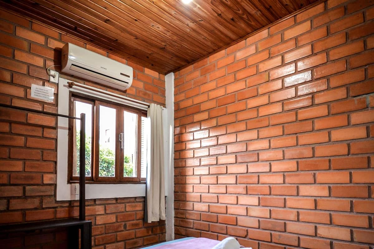 A room with a brick wall, a wooden ceiling, a bed with a pink blanket, and an air conditioning unit.
