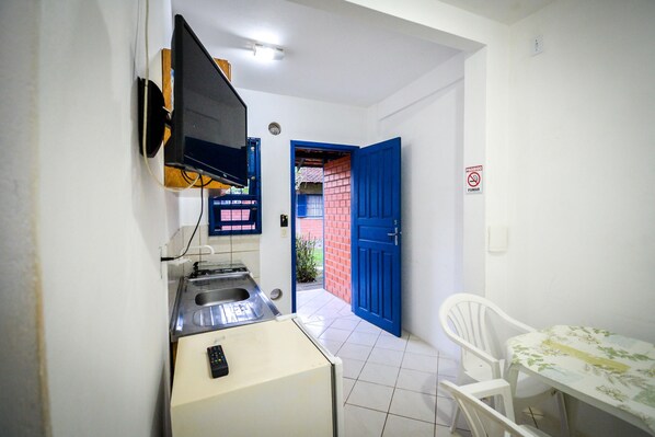 A compact kitchen with a sink, a microwave, and a small refrigerator. There is a blue door leading to an outdoor area with a red brick wall. A white chair and a table with a floral tablecloth are visible in the room. - Pousada Natureza Florianopolis (Florianópolis)