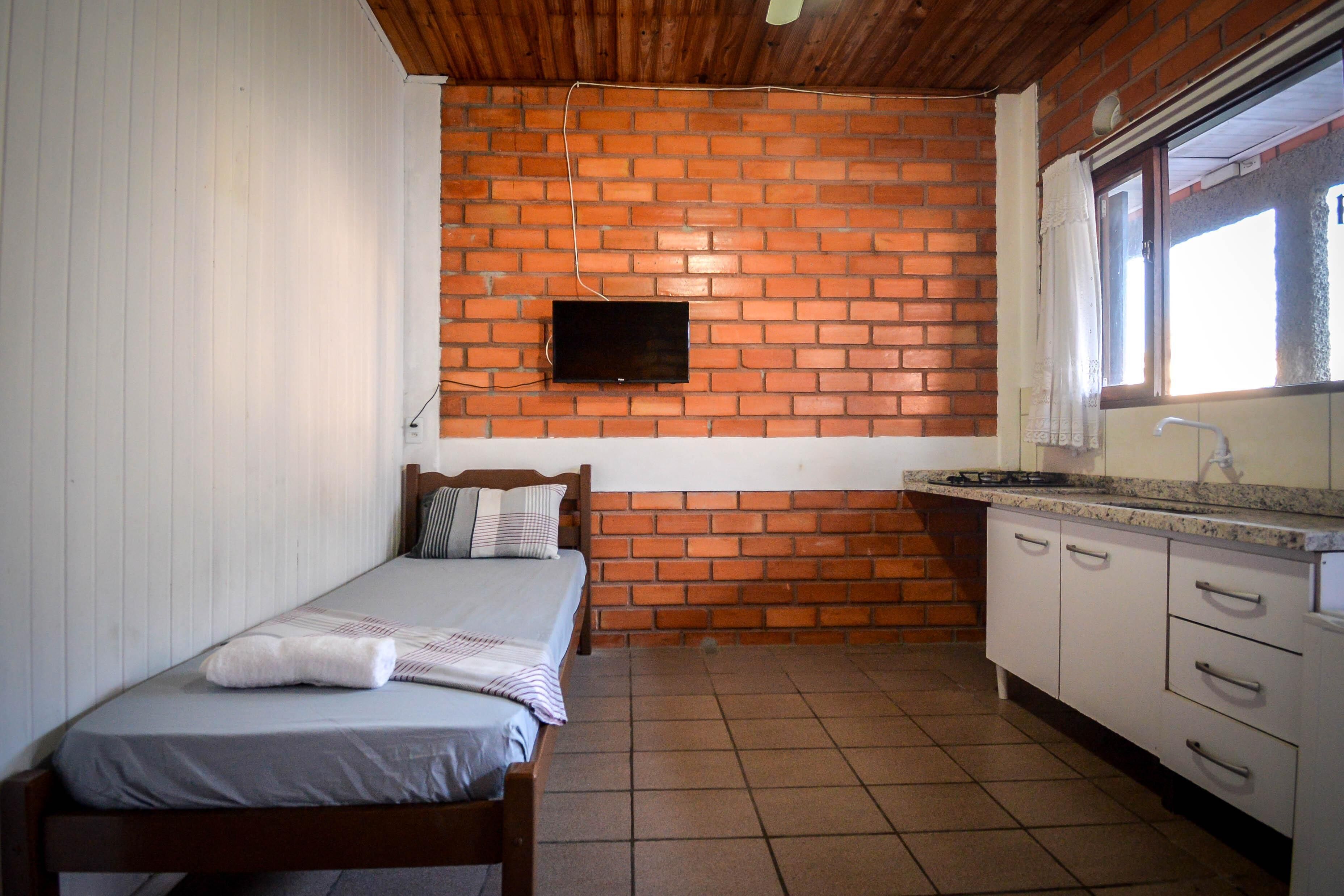 Une petite pièce, de taille modeste, comprenant un lit simple, un mur en briques, un téléviseur fixé au mur et un coin cuisine avec des meubles blancs et un plan de travail.