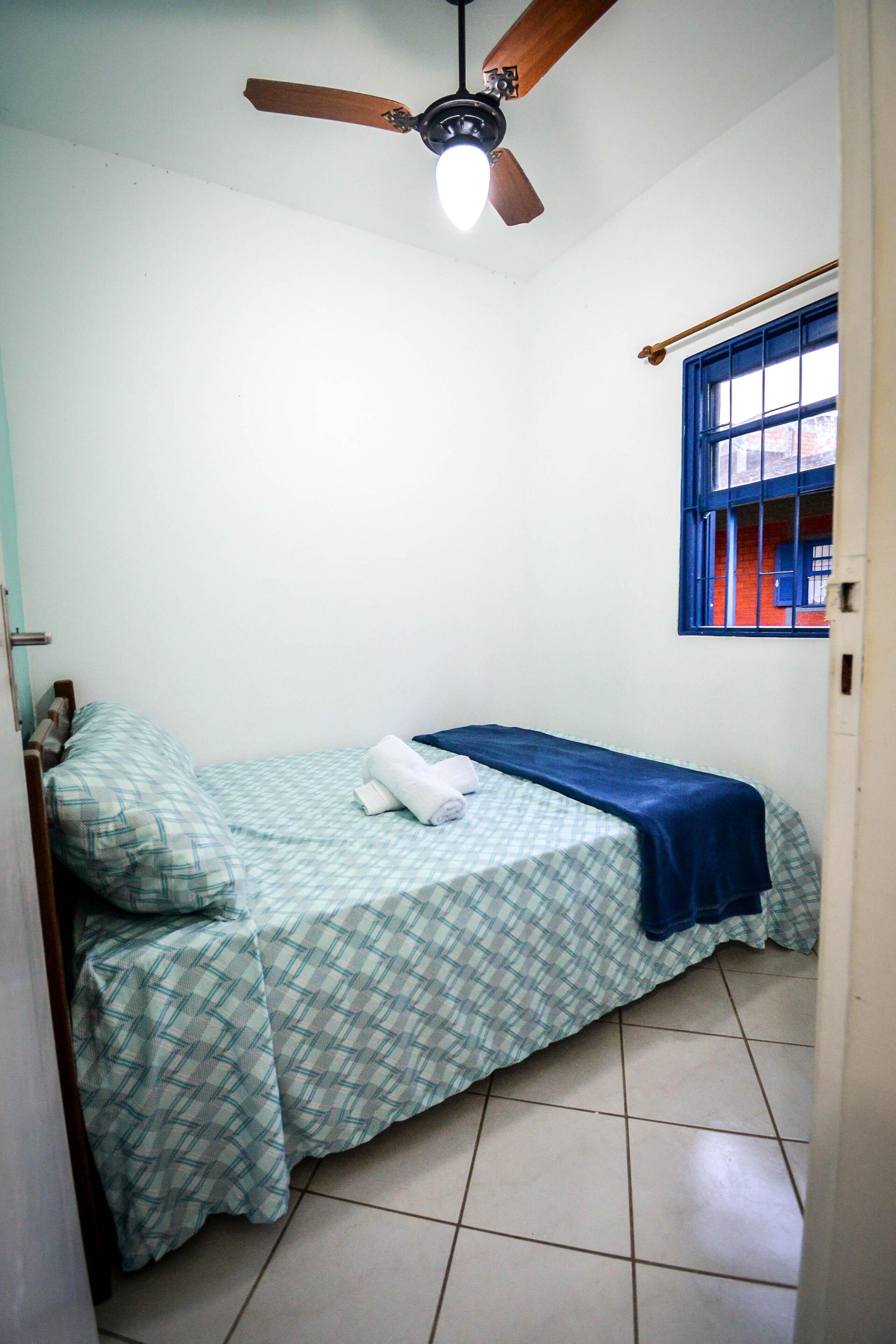 A small, clean room with a single bed, a ceiling fan, and a window with blue bars.