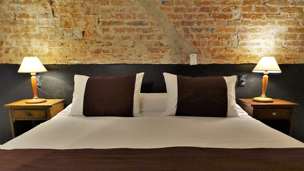 A bed with white linens and two brown pillows, flanked by wooden nightstands with lamps, against a brick wall. - Hosteria del Pescador (Punta del Diablo)