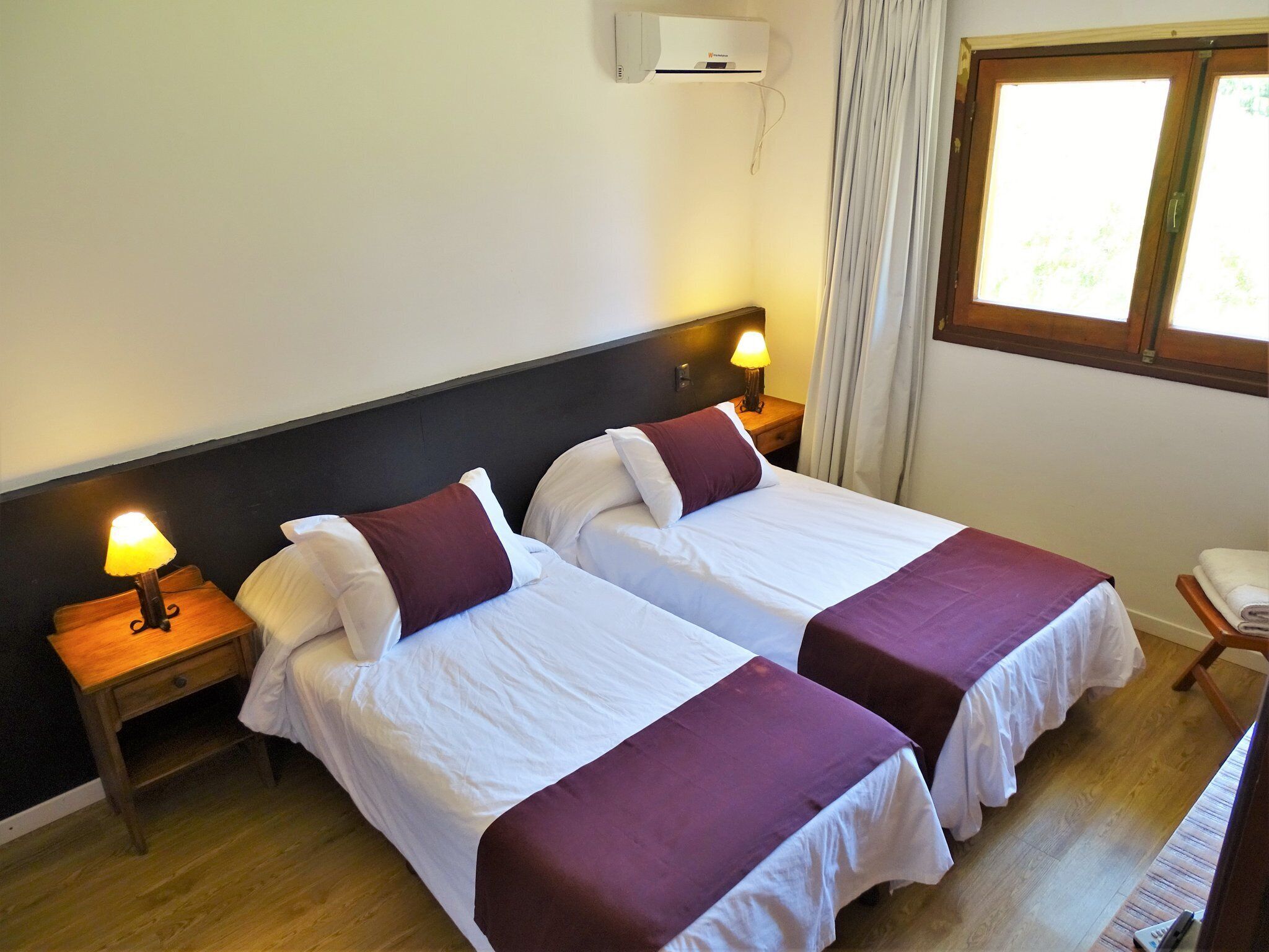 Ein Hotelzimmer mit zwei Betten, jeweils mit einer weißen und violetten Tagesdecke, einem Nachttisch mit Lampe und einer Klimaanlage an der Wand.