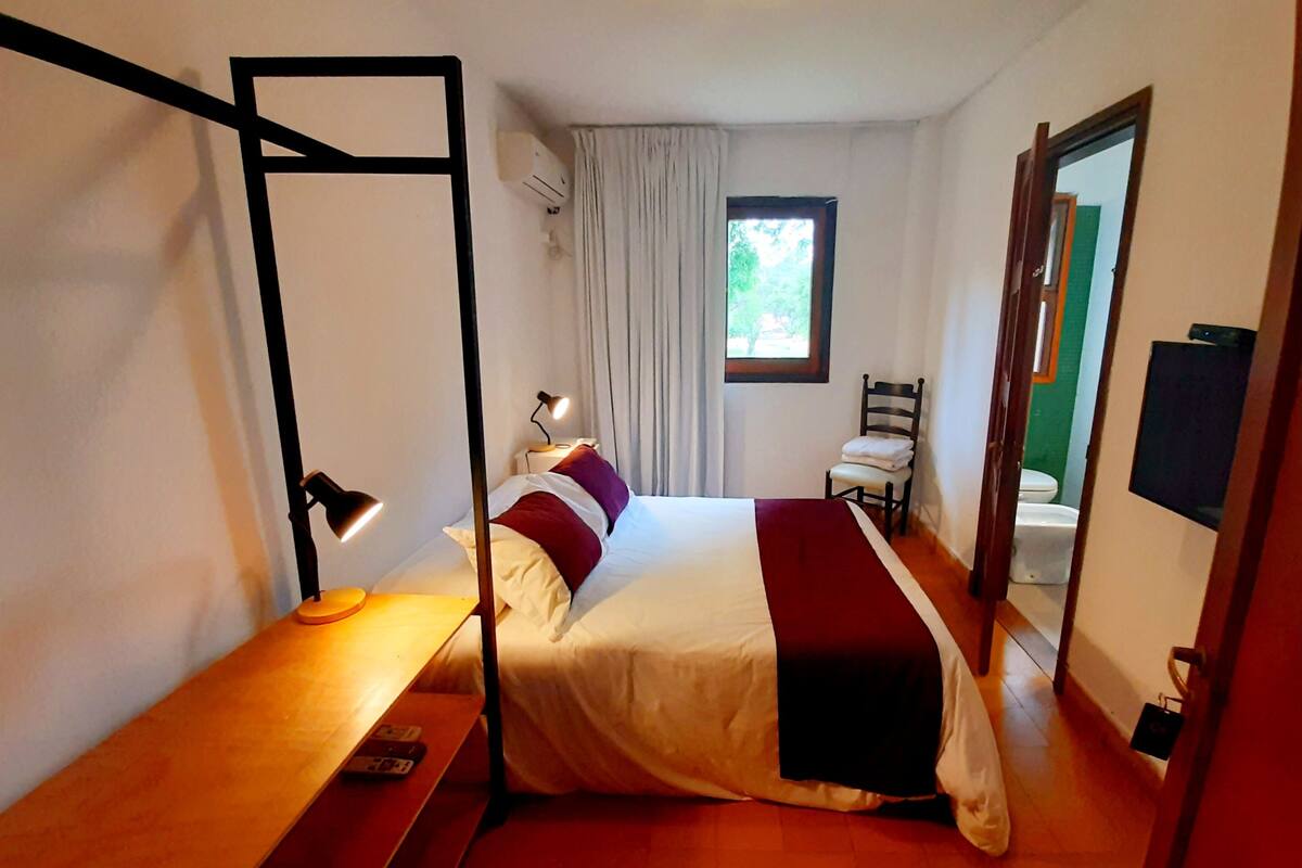 Une chambre d’hôtel de taille réduite, comprenant un lit, une table de chevet, une lampe et une fenêtre avec des rideaux.