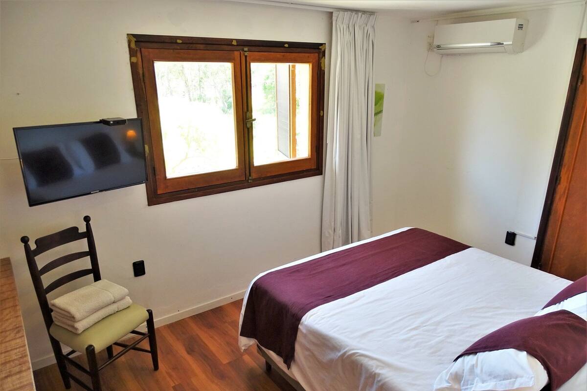 Une chambre à coucher comprenant un lit, une chaise, un téléviseur fixé au mur et une fenêtre avec des rideaux.