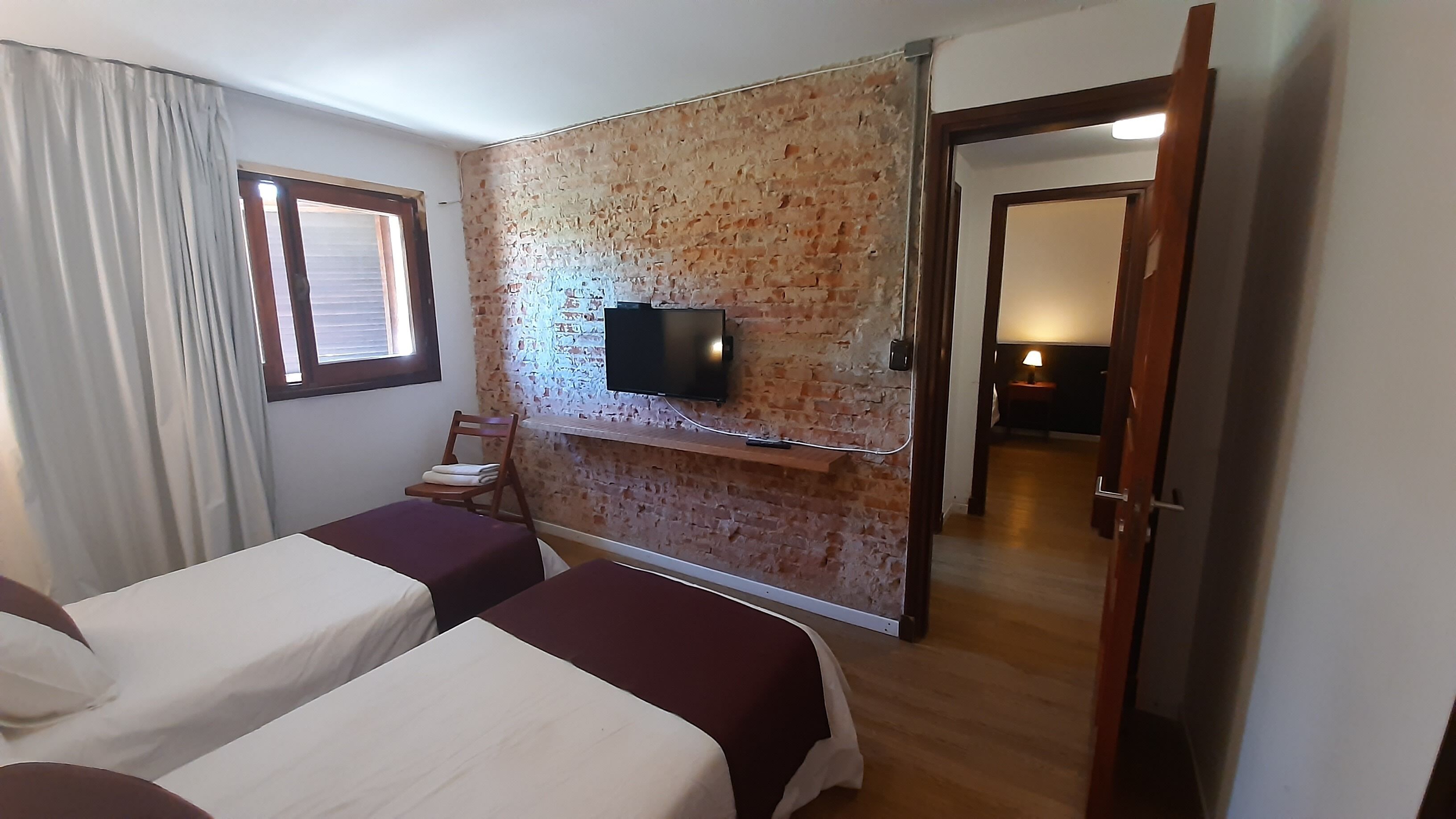Habitación de hotel con dos camas, una pared de ladrillo, un televisor montado en la pared y una puerta que da a otra habitación.