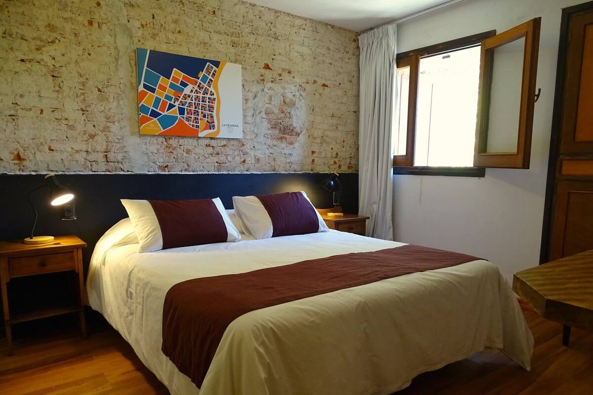 Une chambre avec un grand lit, une tête de lit en bois, deux tables de chevet avec des lampes, une carte accrochée au mur et une fenêtre avec des rideaux.