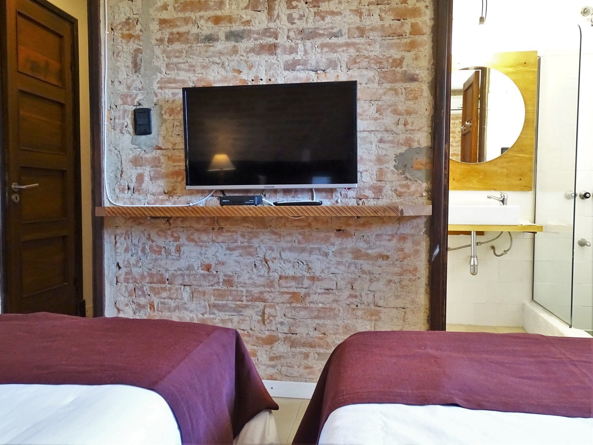 A room with a brick wall, a mounted TV, a wooden shelf, a bathroom with a mirror, and two beds with purple bedding.