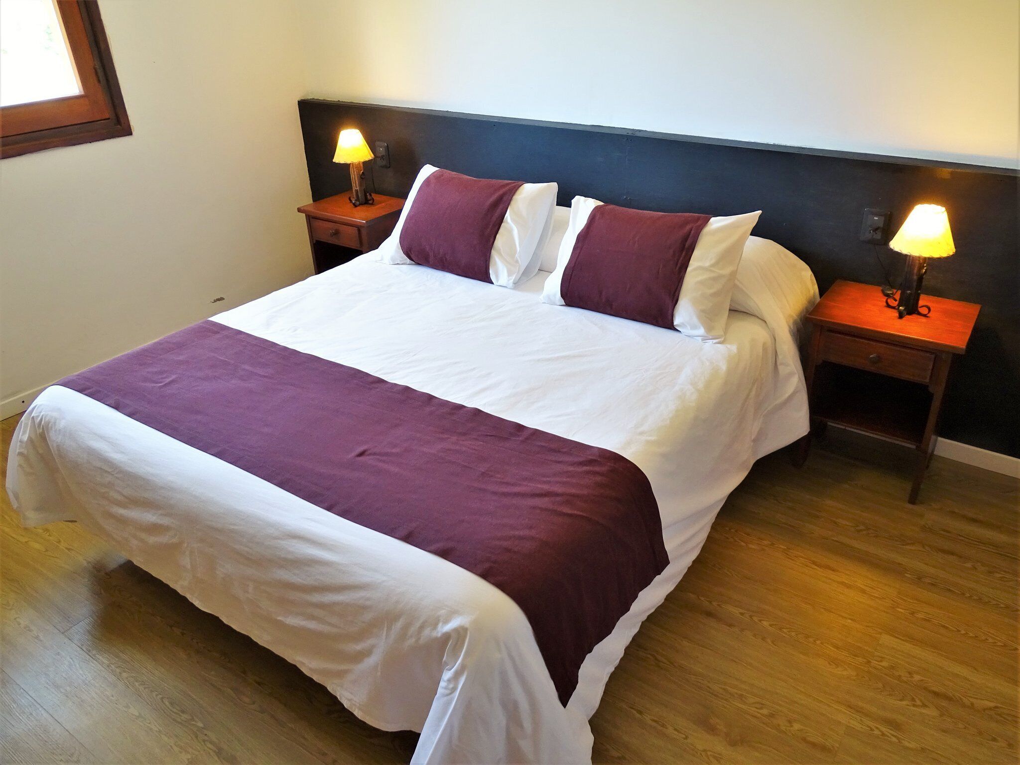 Een net opgemaakt bed met een wit en paars deken, twee bijpassende kussens en twee bedlampjes op houten nachtkastjes.