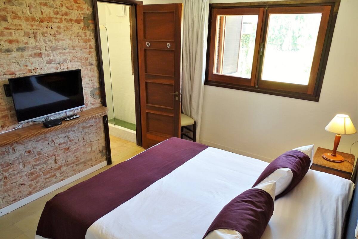 Une chambre d’hôtel avec un mur en briques, une télévision, un lit recouvert d’une courtepointe violette et blanche, une fenêtre avec des rideaux et une table de chevet avec une lampe.
