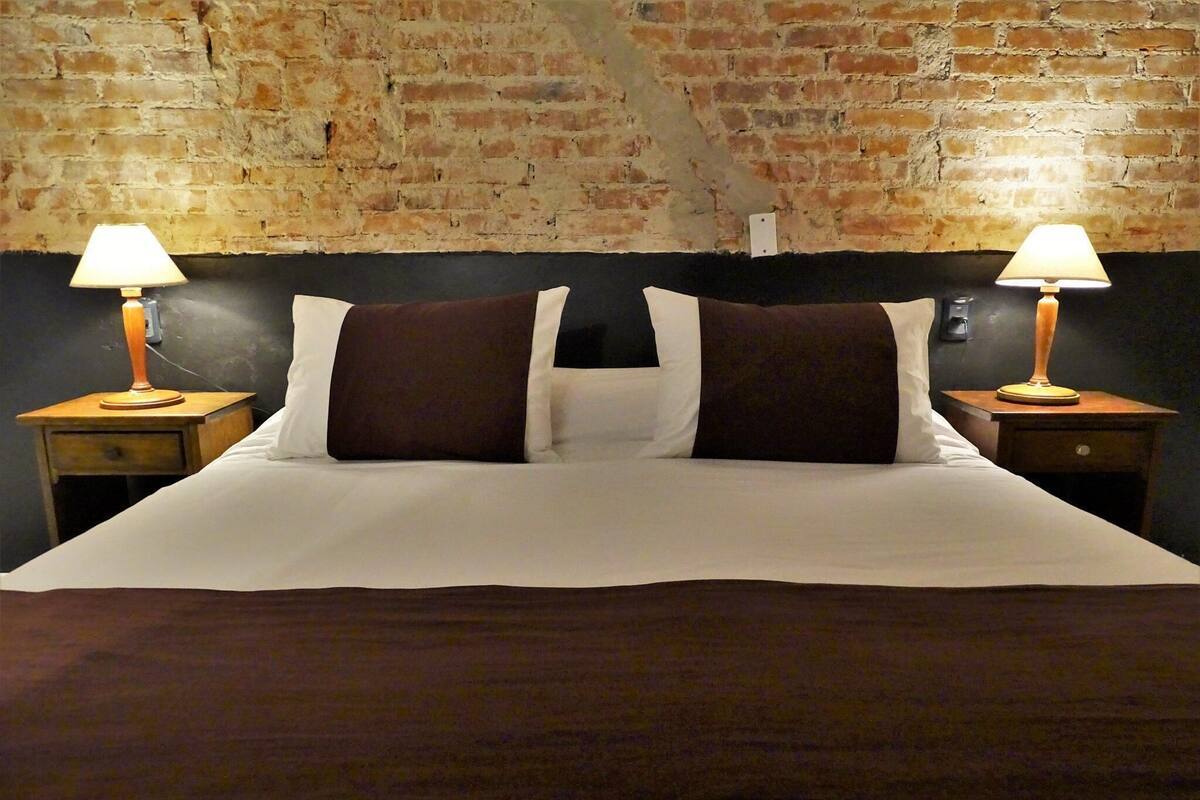 Un lit avec du linge de lit blanc et deux oreillers marron, flanqué de deux tables de chevet en bois avec des lampes, le tout adossé à un mur en briques.