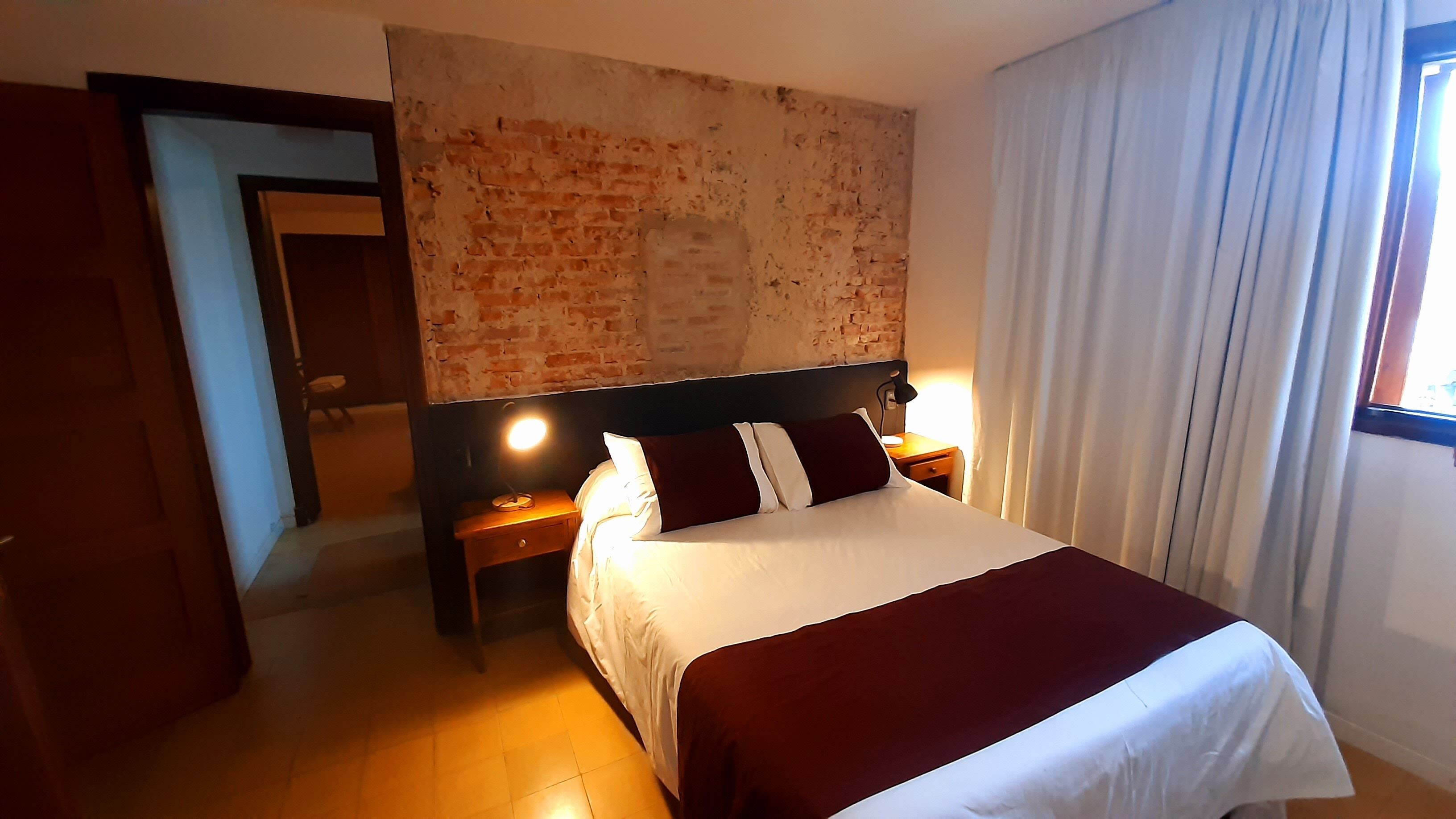 A hotel room with a brick accent wall, a large bed with white and maroon bedding, two bedside tables with lamps, and a window with curtains.