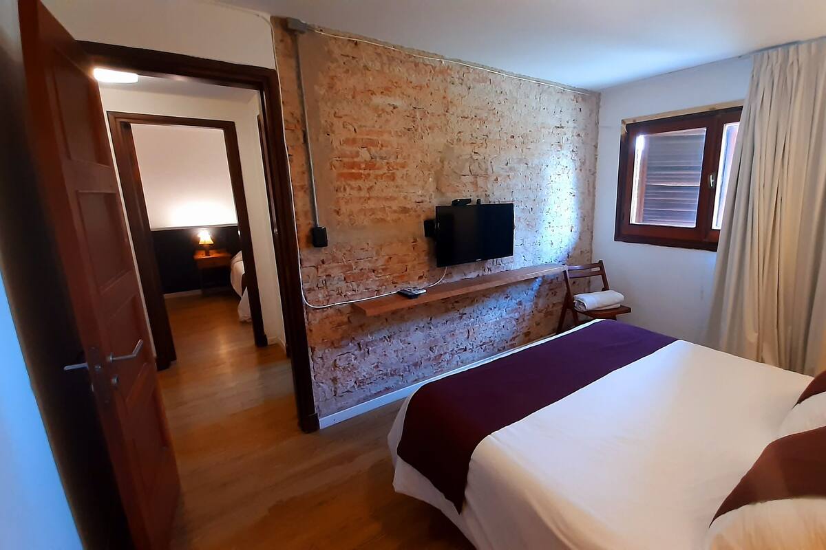 Une chambre d’hôtel avec un mur en briques, un téléviseur fixé au mur, un lit recouvert d’une couverture violette et une fenêtre avec des rideaux.