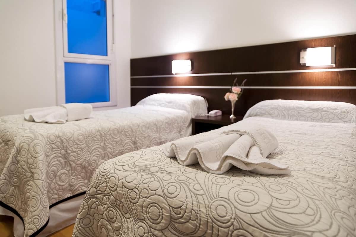 A hotel room with two beds, each with a patterned bedspread, a bedside table with a lamp, and a window with a blue tint.
