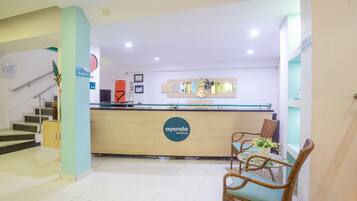 Zona de recepción de un hotel con mostrador, escalera y área de estar con sillas de mimbre.