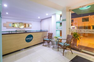 Zona de recepción de hotel con un mostrador de madera, sillas de mimbre, una mesita redonda con un jarrón de flores y una puerta de cristal que da al exterior.