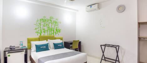 A hotel room with a bed, a desk, a chair, a standing fan, an air conditioning unit, and a wall clock.