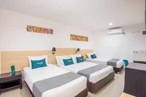 Habitación de hotel con tres camas, cada una con cubrecama gris y blanco y almohadas color turquesa. Se observa un cabecero de madera, una mesita de noche y un escritorio con una silla.