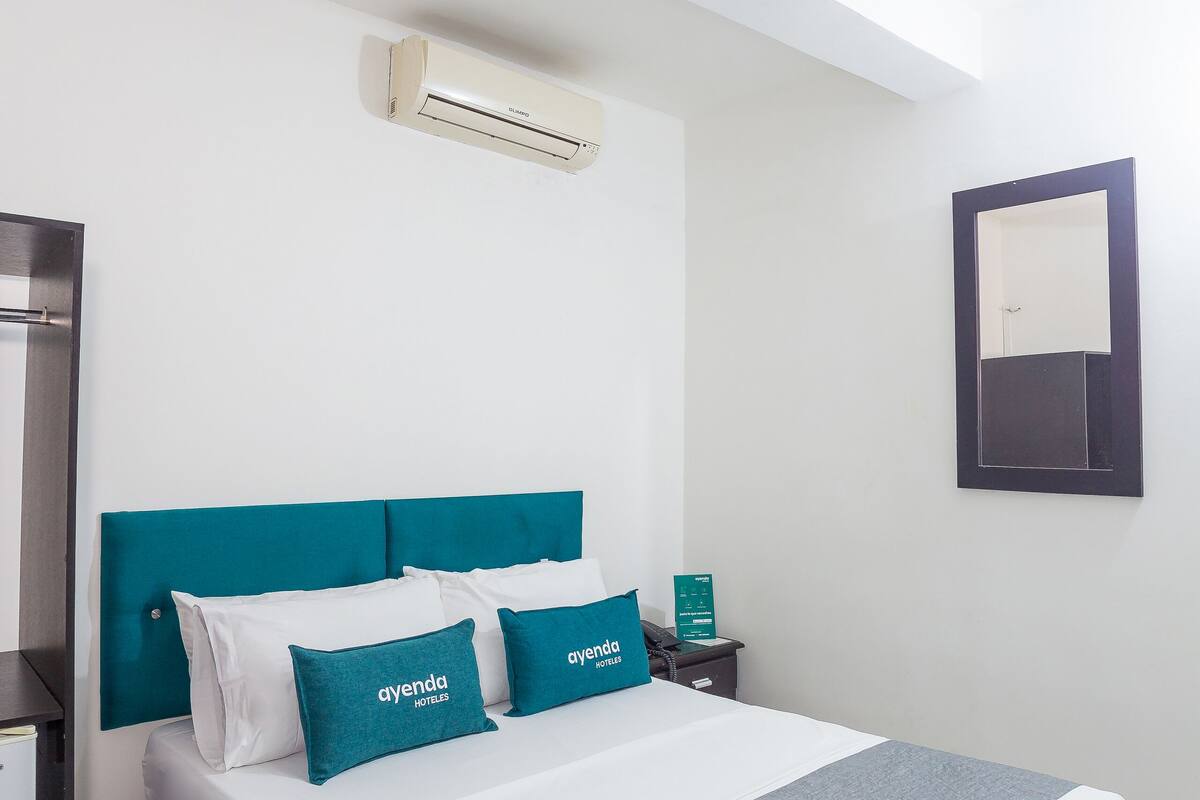 Une chambre d’hôtel moderne avec un grand lit, une literie aux couleurs turquoise et blanche, un système de climatisation intégré, une table de chevet et une petite table de nuit.