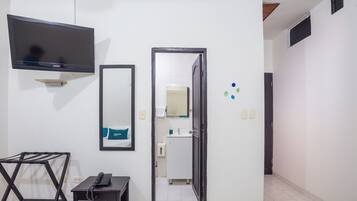 A hotel room with a flat-screen TV mounted on the wall, a small bedside table with a phone, a towel rack, and a door leading to a bathroom with a mirror and sink.