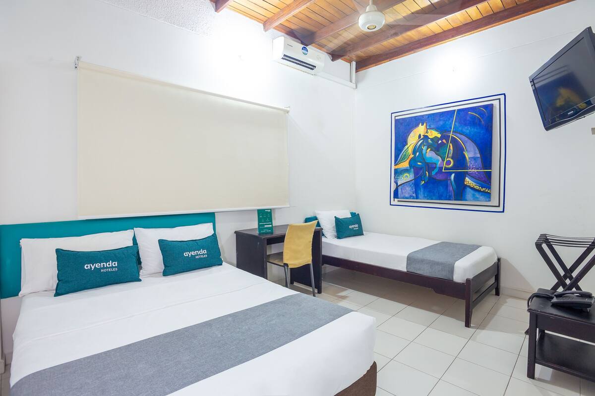 Une chambre d’hôtel avec un grand lit, deux petits lits, un bureau, une chaise, une télévision et un tableau au mur.