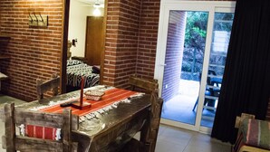 A dining area with a brick wall, a wooden table, and chairs. There is a sliding glass door leading to an outdoor area with a bench and greenery. - Agostina´s (Valeria del Mar)