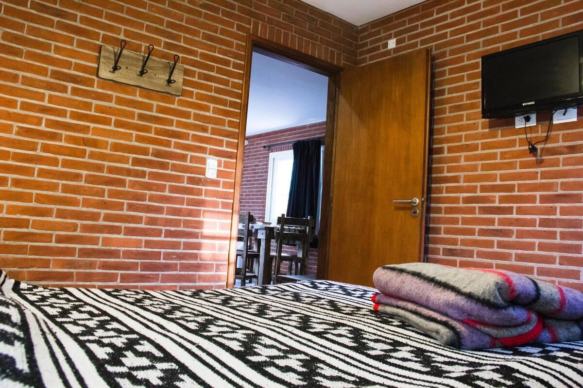 Una stanza con parete in mattoni, un letto con coperta a fantasia, una televisione fissata al muro e una porta che conduce in un'altra stanza.