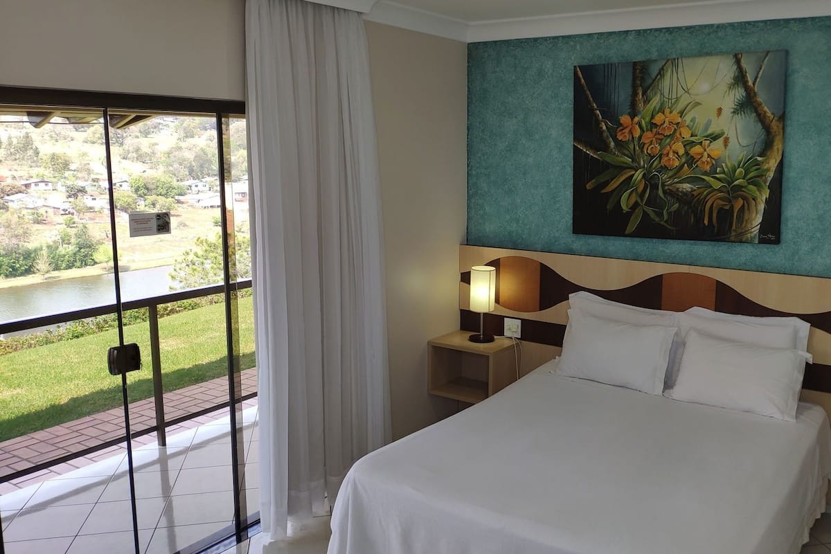 Un cuarto de hotel con una cama, una mesita de noche con lámpara, un cuadro en la pared y vista a un jardín por la ventana.