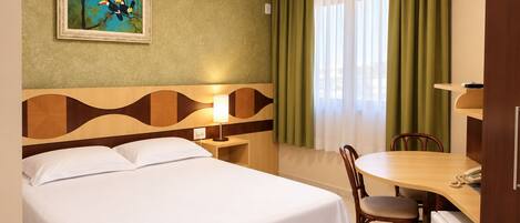 A hotel room with a bed, a desk with a chair, a painting on the wall, and a window with curtains.