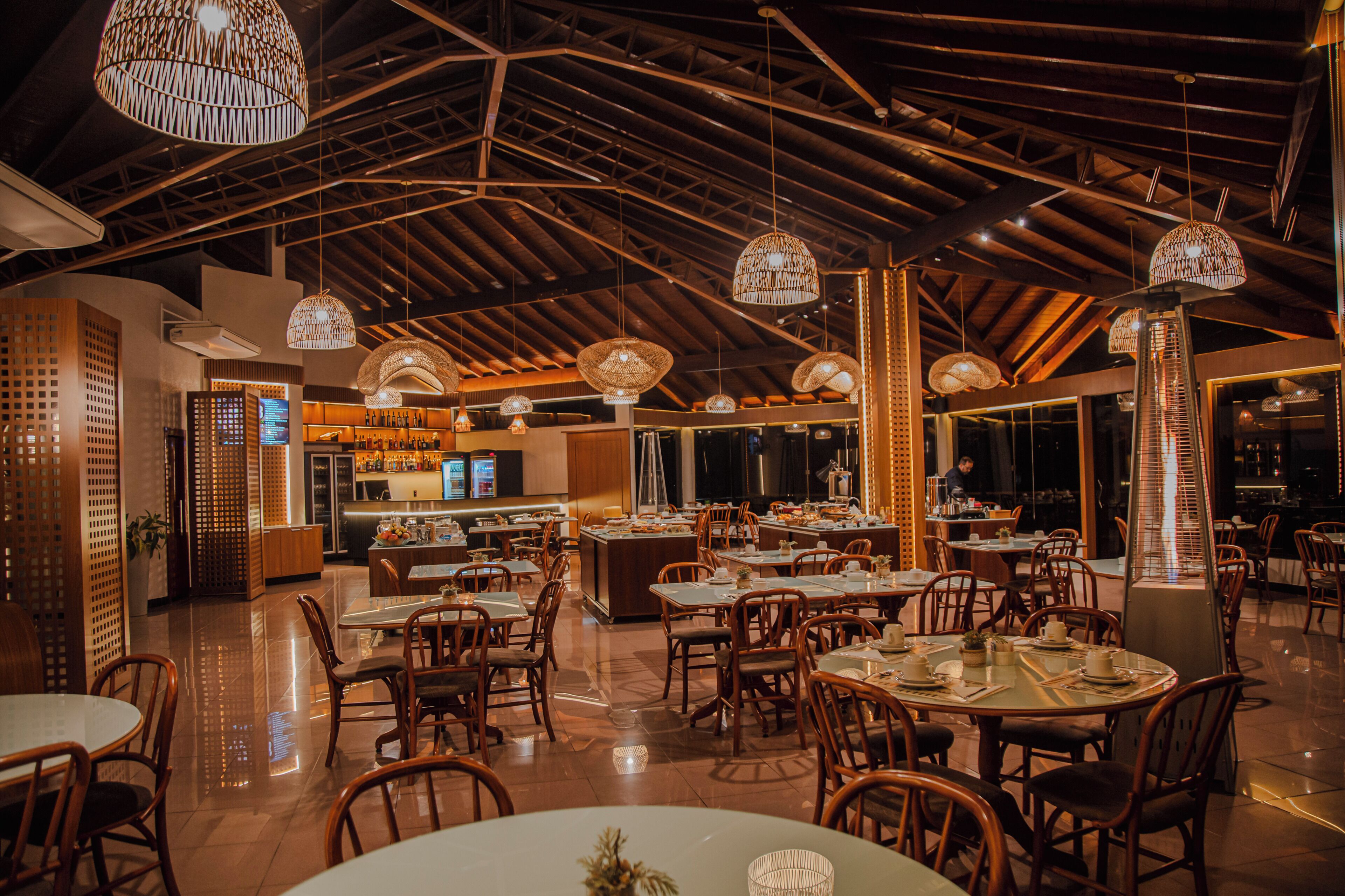 A restaurant with high ceilings, wooden beams, and hanging wicker lamps. Tables are set with chairs, and a bar is visible in the background.