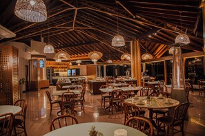 A restaurant with high ceilings, wooden beams, and hanging wicker lamps. Tables are set with chairs, and a bar is visible in the background.