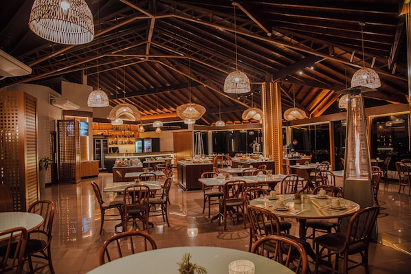 A restaurant with high ceilings, wooden beams, and hanging wicker lamps. Tables are set with chairs, and a bar is visible in the background.