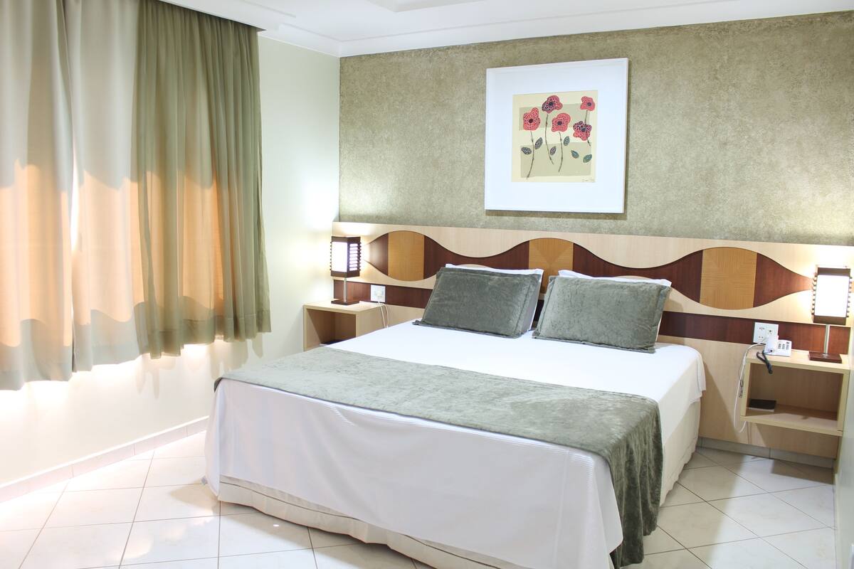 Una habitación de hotel con una cama grande, dos mesitas de noche, una obra de arte en la pared y una ventana con cortinas.