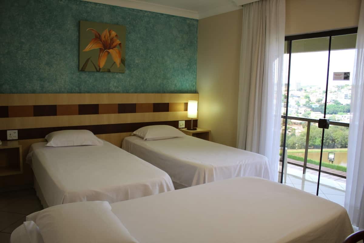 Un cuarto de hotel con dos camas individuales, una mesita de noche con lámpara, un cuadro en la pared y una ventana con vista a la ciudad.