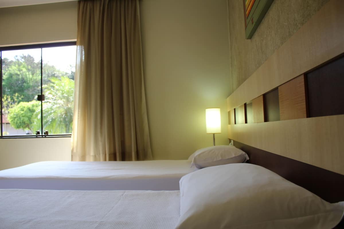 Habitación de hotel con dos camas, una ventana con cortinas, una lámpara de pared y vista a zonas verdes.
