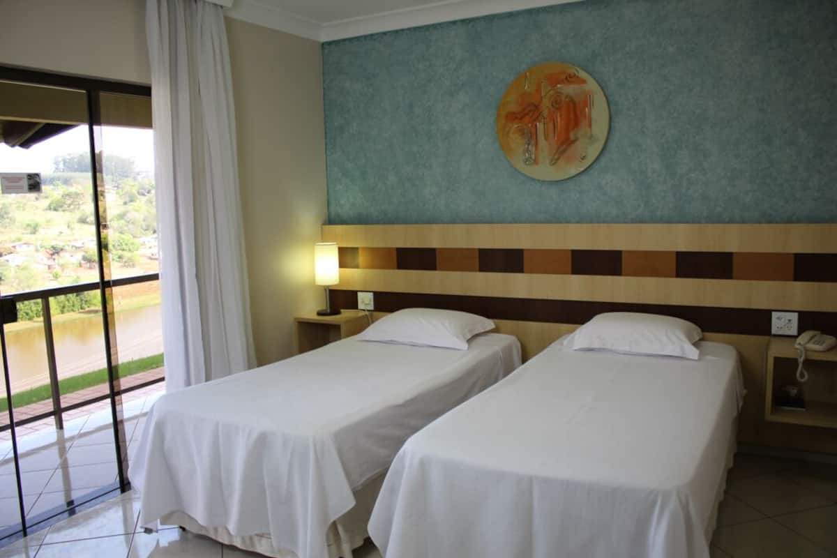 Una habitación de hotel con dos camas individuales, un ventanal con vistas a zonas verdes, un cuadro en la pared y una mesita de noche con una lámpara.