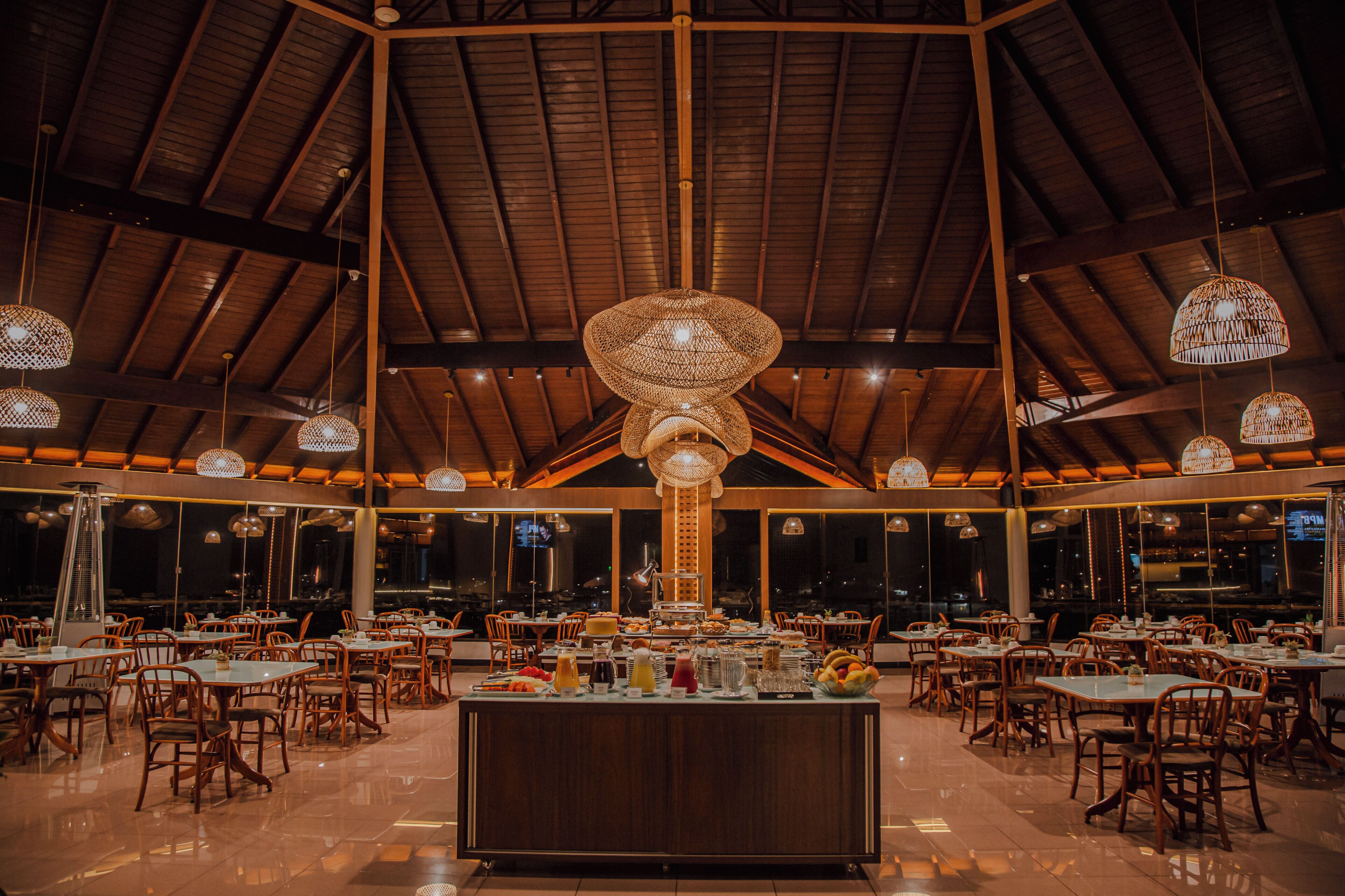 A spacious restaurant with high ceilings and multiple hanging light fixtures. Tables are arranged around a central bar area.