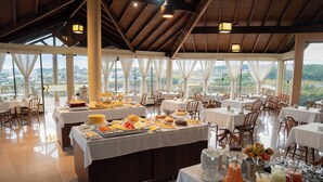 Un buffet proposant une variété de plats et de boissons, disposés sur des tables recouvertes de nappes blanches, dans une pièce spacieuse dotée de grandes fenêtres et offrant une vue sur le paysage.