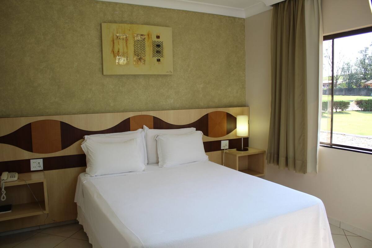 Una habitación de hotel con cama, mesita de noche, lámpara, teléfono y ventana con cortinas.